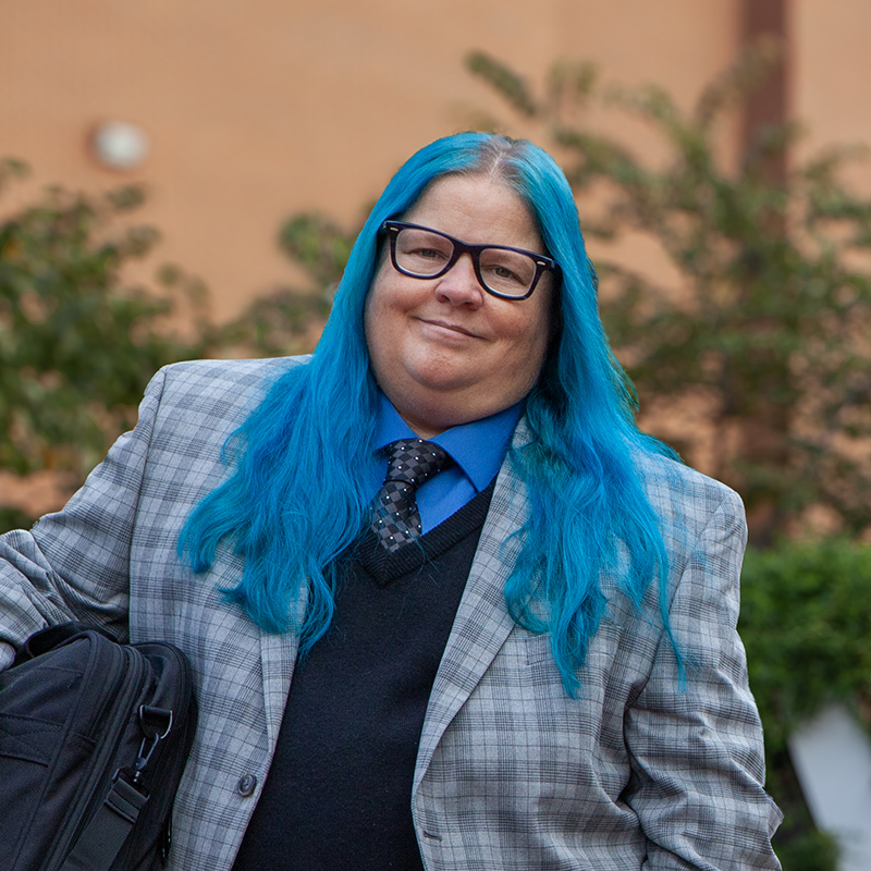 Steph Cull with long blue hair and blue checked jacket smiles at the camera