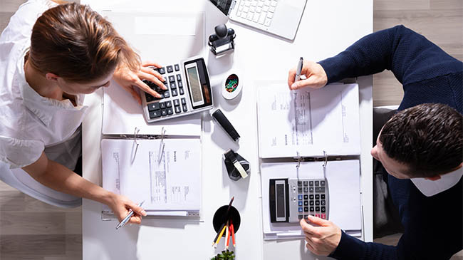 Two individuals sitting at a table, engrossed in calculations, using a calculator and examining papers.