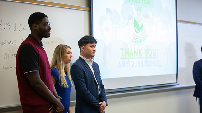 Three students standing in front of a whiteboard presenting a business plan.