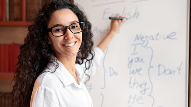 A woman in glasses writing on a whiteboard in a classroom.