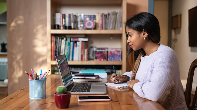 A woman sitting at a table with a laptop and notebook, working diligently.