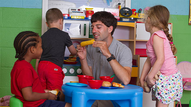 A man and two children happily playing with blocks in a classroom.