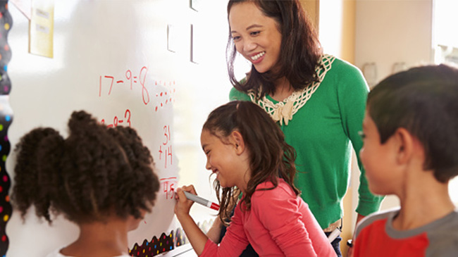 A teacher writing on a whiteboard with children, engaged in a classroom activity.