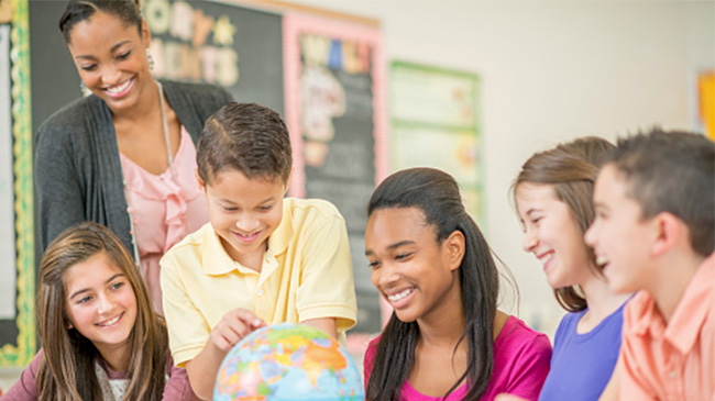 A diverse group of children sitting at desks in a classroom, listening attentively to the teacher.