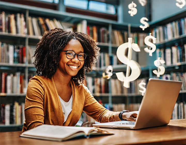 a woman sitting in a library smiles at her laptop. The laptop has dollar signs flying out of the screen.