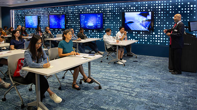 A classroom with people sitting at desks and a man giving a presentation.