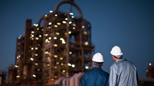 . Two men in hard hats standing in front of an oil refinery, discussing safety measures at the industrial site.