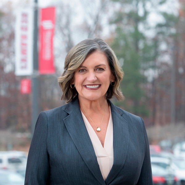 Professional headshot of a woman in a dark gray business suit and cream-colored blouse. She has shoulder-length wavy gray-blonde hair, is wearing small hoop earrings and a pearl pendant necklace, and is smiling warmly at the camera. The background shows red banners and trees on the Reynolds Parham Road campus.